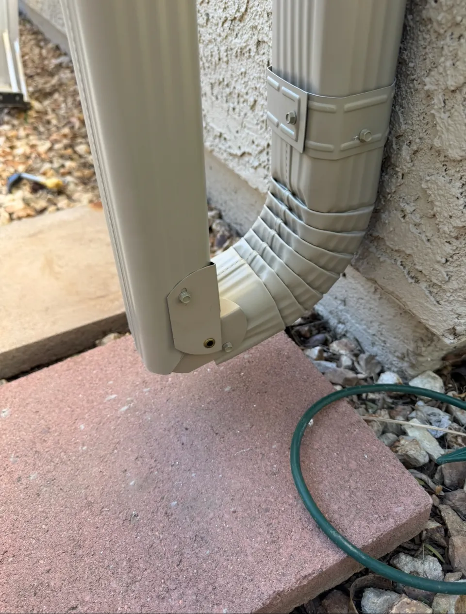 A downspout elbow discharging onto a splash block beside pavers and gravel